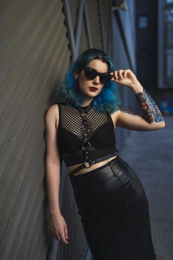 A European female with tattoo and blue hair wearing sunglasses and sexy black dress posing at the side of a building