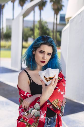 A vertical shot of a cool young lady with blue hair holding a mask in her hand posing in the street