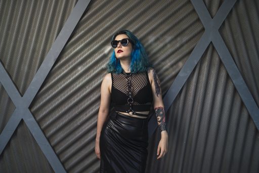 A European female with tattoo and blue hair wearing sunglasses and sexy black dress posing at the side of a building