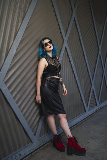 A cool young lady with blue hair, tattoos, and sunglasses, wearing a crop top, leather pencil skirt and red shoes leaning on a wall