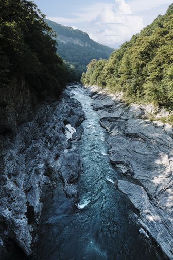 A vertical shot of a river in the middle of cliffs with trees under a blue sky at daytime