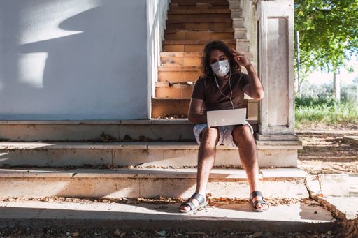 A young man wearing a face mask while sitting on an old outdoor stairs and communicating though his smartphone
