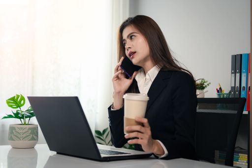An Asian female professional in a formal business suit is talking on her phone while holding a cup of freshly brewed coffee.