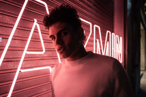 A Hispanic male wearing street-style fashion clothes posing in front of a pink neon sign