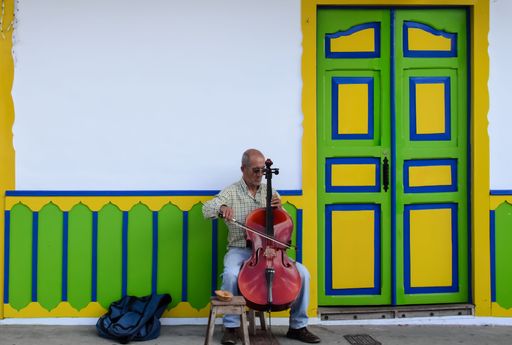 A male street performer playing cello in front of a colorful door and wall building