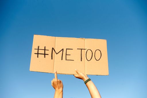 A cardboard sign saying hashtag Me Too held by two hands on clear blue sky background