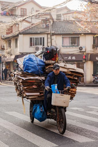 A vertical shot of a man riding a bicycle full of cardboard sheets and a metal basket on a top of them