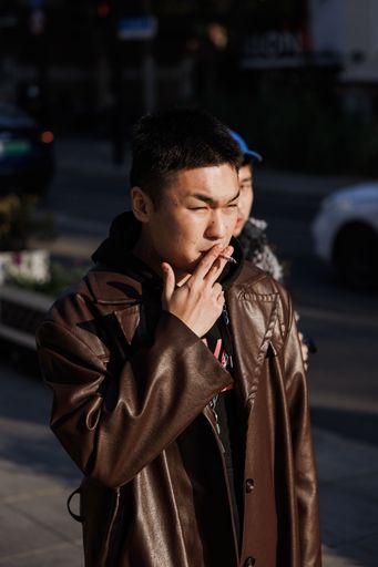 A vertical shot of a cool young male in a brown leather trench coat smoking in the streets of Shanghai