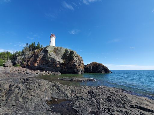 A low angle of a lighthouse on top of a rocky hill covered with green trees against a sea on a sunny day