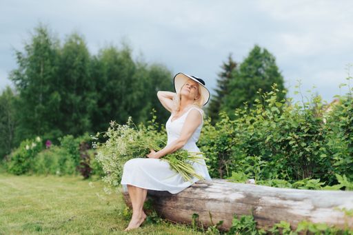 A beautiful blonde woman in a white dress and a hat holding flowers and sitting on a tree trunk