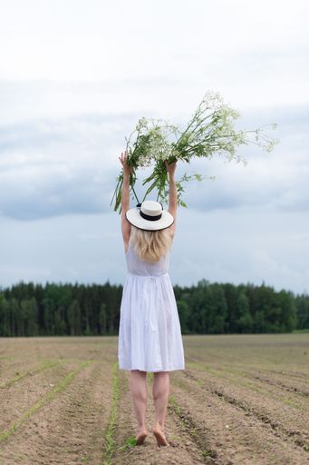A vertical shot of a beautiful blonde woman wearing a hat and a white dress holding flowers in a field