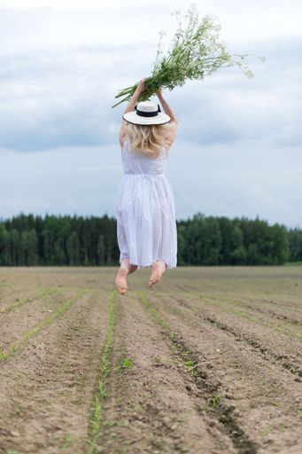 A vertical shot of a beautiful woman in a white dress jumping around in a field holding flowers