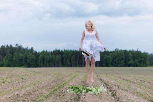 A beautiful blonde woman in a white dress jumping around in a field with green trees in the background