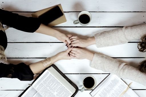 An overhead shot of females holding hands over the table with coffees and open bibles