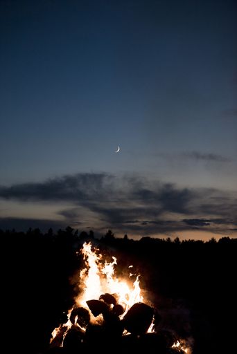 A vertical shot of a campfire with a visible moon in a dark blue sky