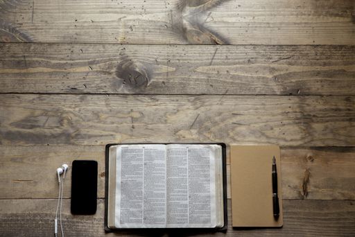 An overhead shot of a opened bible in the middle of a notebook and a smartphone on a wooden table