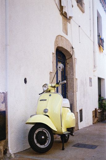 A vertical shot of Italian yellow Vespa scooter parked in front of a white building