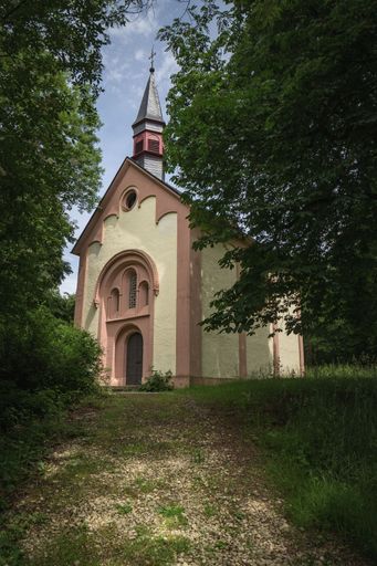 A vertical shot of a little Christian chapel surrounded by green trees under a blue sky, Trier, Germany