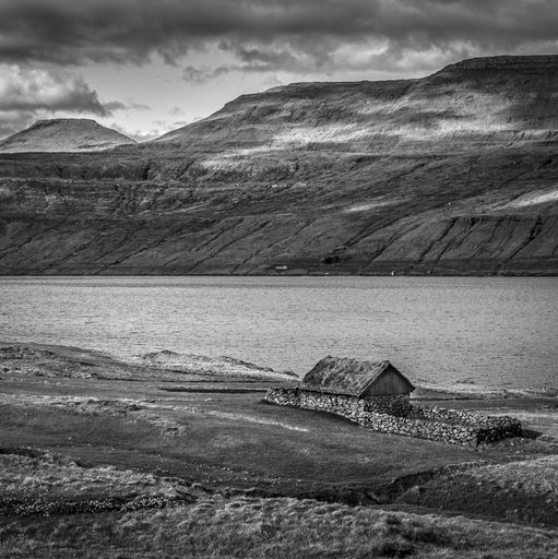 A vertical shot of a house near the river with mountains in the distance under a cloudy sky in black and white