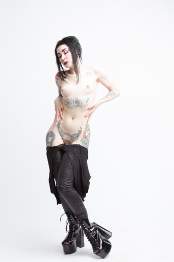 A vertical shot of a half-naked model covered in tattoos wearing tight pants and platform heels posing for a photoshoot