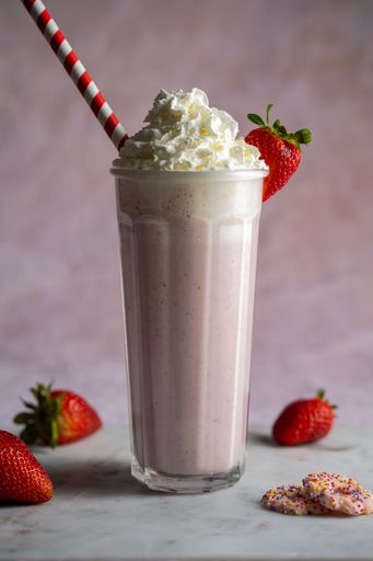 A vertical shot of a delicious fresh strawberry milkshake with a red and white straw in it and sprinkled cookies around the glass