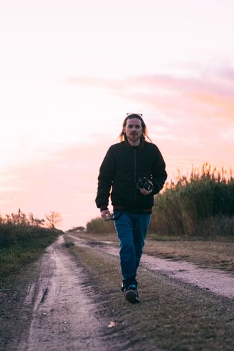 A vertical shot of Caucasian man walking through road surrounded by growing bushes during sunset