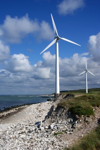 A vertical shot of windmill turbines standing  near a shore in a sunny day against blue sky with white clouds