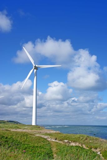A vertical shot of windmill turbine standing on a shore in a sunny day with a blue sky and abundant white clouds