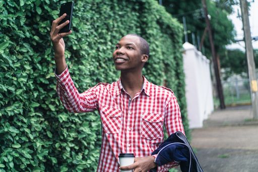 A young African American male in a red and white chequered shirt taking a selfie in a park