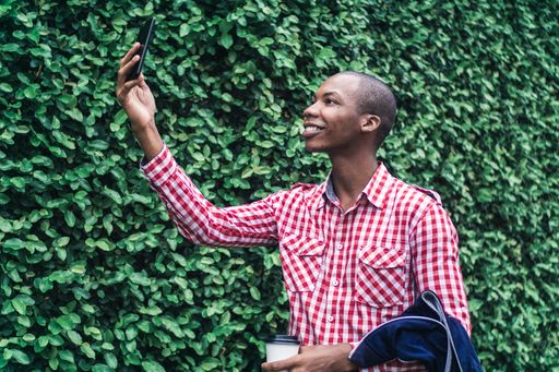 A young African American male in a red and white chequered shirt taking a selfie in a park