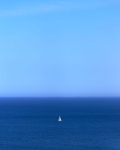A vertical shot of a sailboat on the endless horizon of the sea, shades of blue