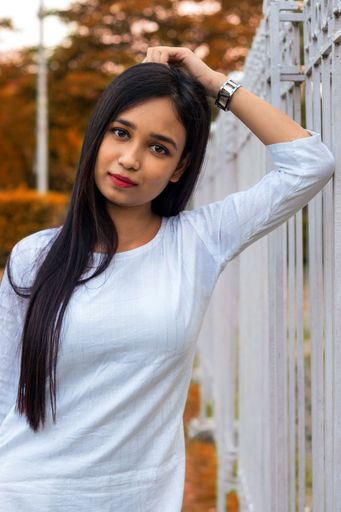 A vertical portrait of a young, Indian woman with white, traditional Kurti, leaning on the fence and looking at the camera