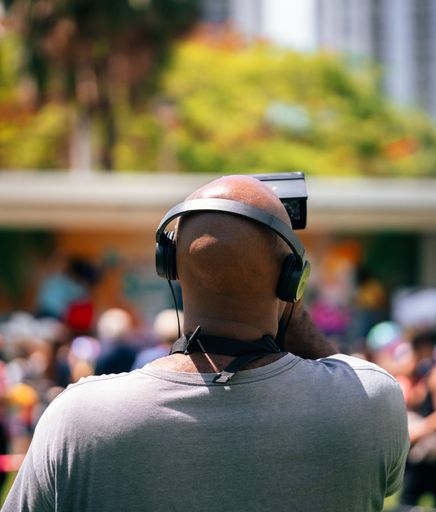 A rear, vertical shot of a cameraman with headphones filming a public event