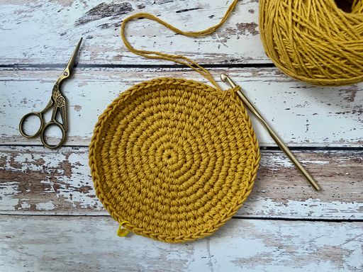 An unfinished crochet round placemat on a wooden surface and a scissor on its side