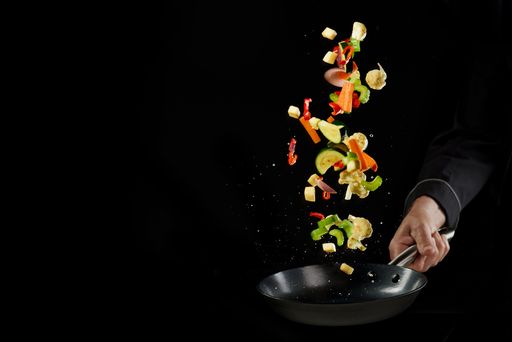 The chef's hand throwing fresh delicious assorted vegetables on frying pan against a black background