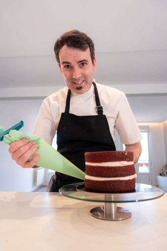 A man cooking a red velvet cake in the kitchen at home, decorating the triple sponge base of the cake with a piping bag assembled with cream