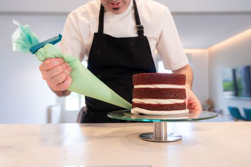 Hands of a man cooking a red velvet cake at home, covering the edges of the triple sponge cake base with the pastry bag filled with cream