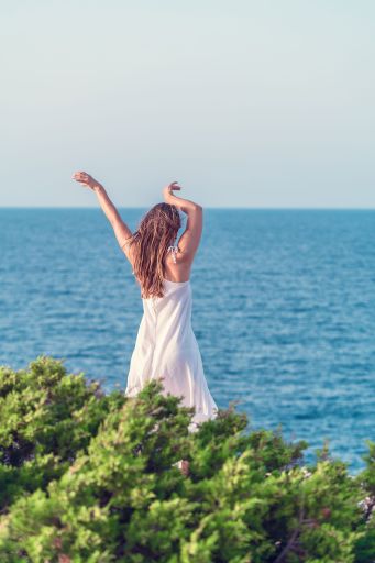 Woman with white dress standing in front of beach with her hands raised and looking into a wide sea view. Concept of vacation and tranquility.