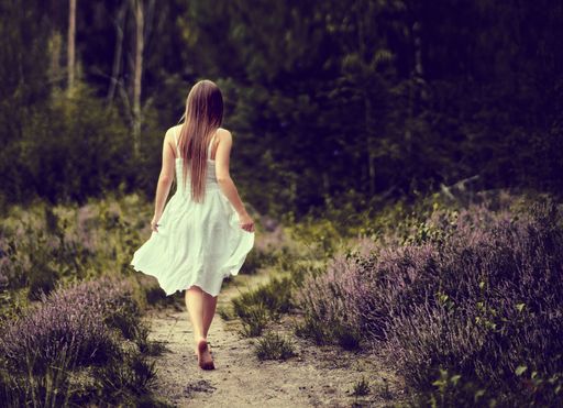 A back view of female in white dress walking on footpath surrounded by blooming trees in forest