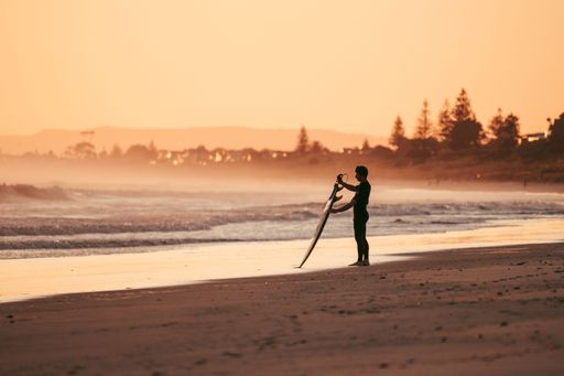 A side view of a male surfer with a board standing on the beach during sunset