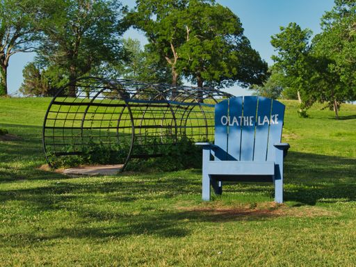 A blue wooden lawn chair near the splash pad in the green field at Olathe Lake