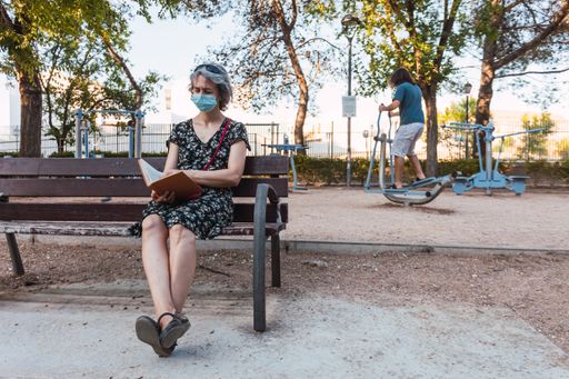 A Caucasian female sitting on a bench of a park reading a book with a Caucasian male training in the background