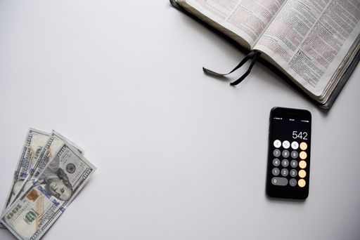 An overhead shot of an open book near a smartphone and hundred dollar bills on the side on a white surface