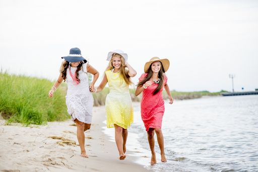Three women wearing colorful dresses and hats running through the beach surrounded by the sea