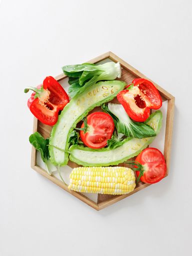 A top view of a wooden board with tomato, pepper, bitter gourd, corn and lettuce on white background