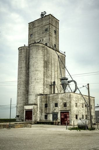 Old, concrete, weathered, grain, elevator with worn red doors, a landmark of the countryside in Midwest Illinois, sits next to rusty railroad tracks.