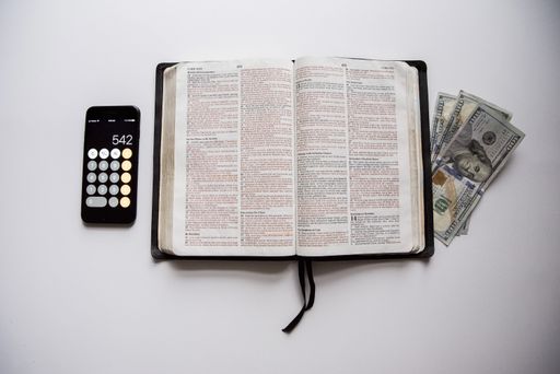 An overhead shot of an open bible in the middle of money and a smartphone on a white surface