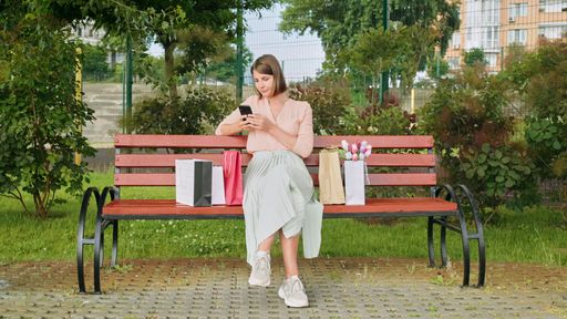 A Caucasian woman sitting with shopping bags on bench and looking at smartphone