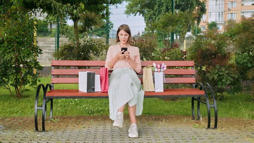 A Caucasian woman sitting with shopping bags on bench and looking at smartphone