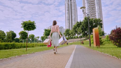 A Caucasian woman holding shopping bags and walking through park in background of buildings
